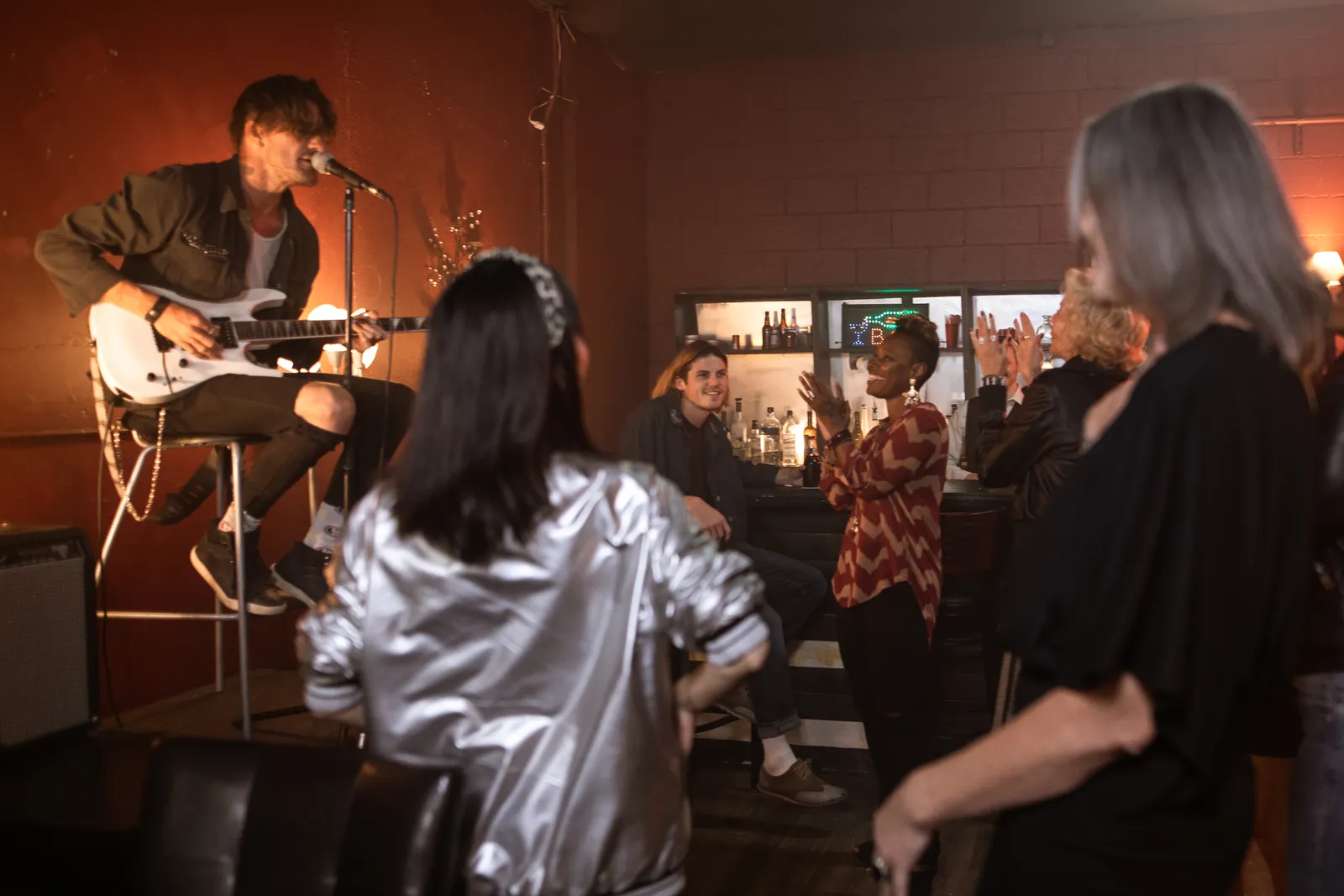 A packed pub back room: a singer with a white guitar plays to a crowd clapping by the bar